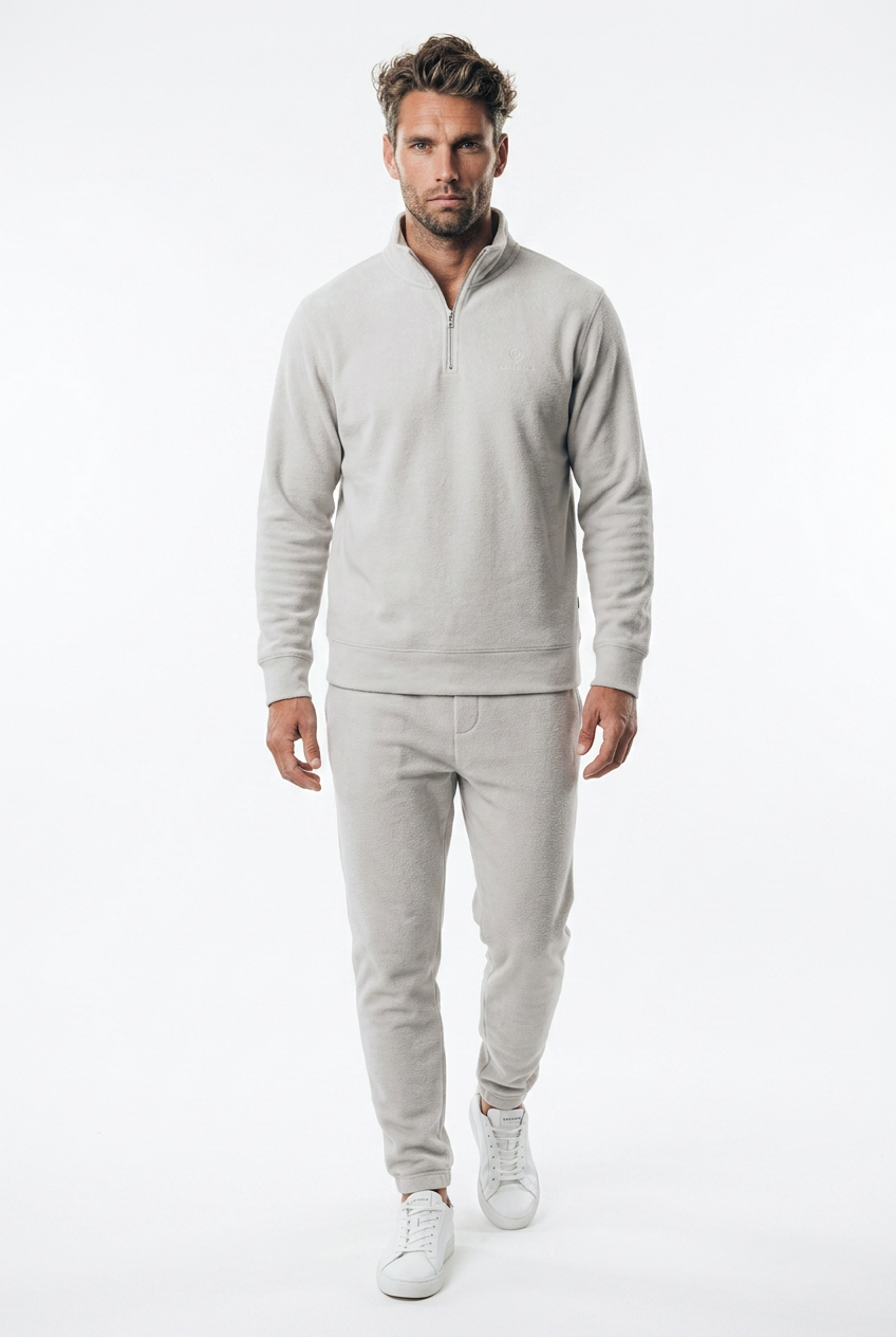 A man modeling a light gray matching two-piece tracksuit, featuring a quarter-zip golf jumper and matching jogger pants, paired with white sneakers against a plain white studio background.