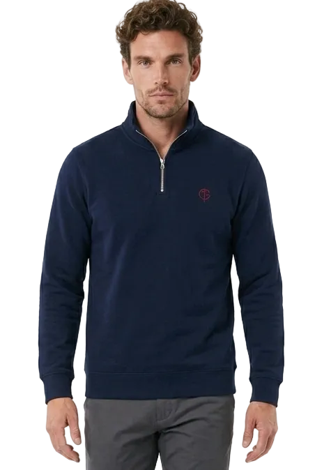 A man modeling a navy blue quarter-zip golf sweater with a small embroidered logo on the chest, paired with dark gray trousers and white sneakers, against a plain white studio background.