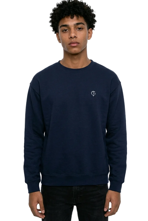 A young man with curly black hair wearing a navy blue crewneck golf sweater with a small white embroidered logo on the left chest, paired with black jeans, against a plain white background.