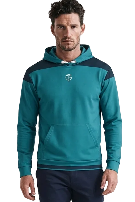 A man modeling a teal and navy blue colorblock golf hoodie with a small white chest logo, paired with navy golf pants and white sneakers, against a plain white studio background.