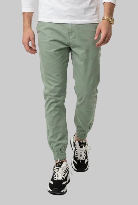 A man modeling a white long-sleeve crewneck t-shirt and sage green slim-fit golf trousers with black and white sneakers, shot against a plain white studio background.